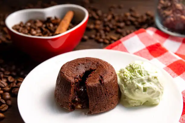 Photo of A portion of chocolate fondant with a scoop of pistachio ice cream on a plate. In the background is a heart-shaped cup filled with coffee beans and a cinnamon stick.