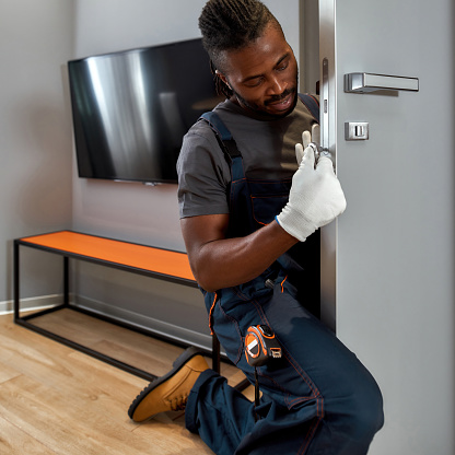 Closeup portrait young African locksmith in uniform, sitting on floor, fixing appartment door lock. Skilled handyman in modern flat interior. Diverse community concept.