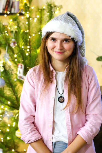 retrato desenfocado de una mujer sonriente joven de 20 a 25 años con sombrero blanco de santa y camisa rosa. posando en el interior con el árbol de navidad de fondo. vertical. navidad. fuera de foco - 20 25 years color image contemporary domestic life fotografías e imágenes de stock