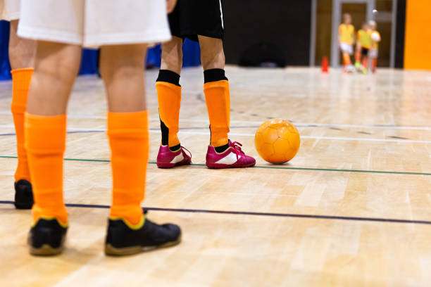 屋内サッカートレーニングの子供たち。子供たちは、木製の床にフットサルを再生します。オレンジのフットサルボールを持つ子供サッカー選手の足。練習クラスで2つの反対の列に立つプレ - futsal indoors soccer ball soccer ストックフォトと画像