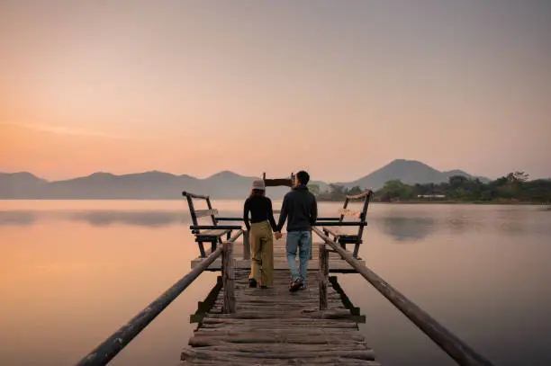 Photo of Young asian couple walking and holding hands on wooden bridge among the reservoir
