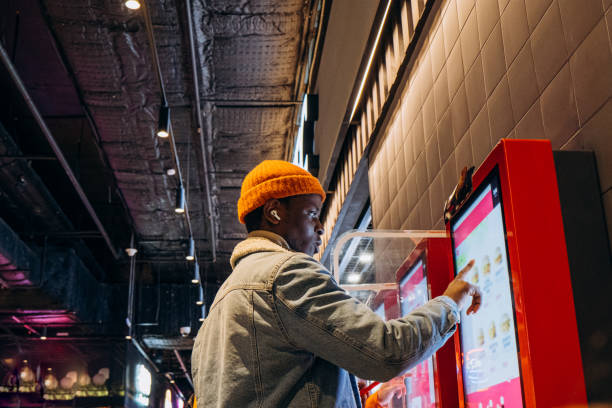 African-American man uses self-service kiosk to order snack Smiling African-American man in warm denim jacket with wireless earphones uses self-service kiosk to order snack in cafe black orange audio stock pictures, royalty-free photos & images