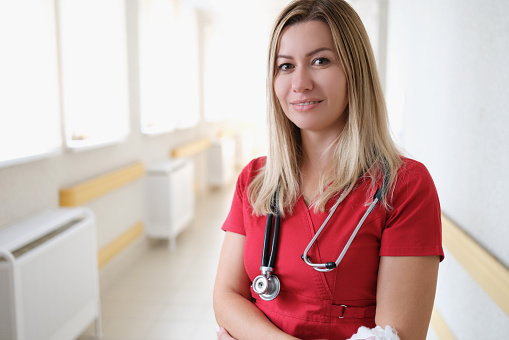 Portrait of young female doctor with stethoscope in red uniform in hospital corridor. Insurance medical assistance concept