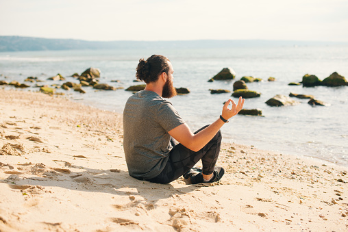 Relaxed bearded sporty man is sitting in the lotus pose on the beach, resting after running.