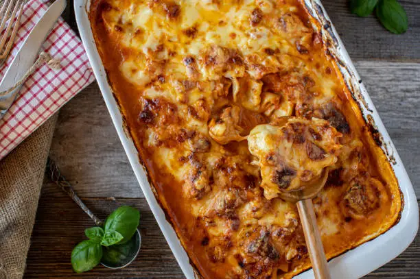 Delicious homemade noodle casserole or noodle gratin cooked with tortellini al forno in a delicious vegetable, tomato sauce and a mozzarella cheese topping. Served in a white baking dish on rustic and wooden table. Closeup and table top view