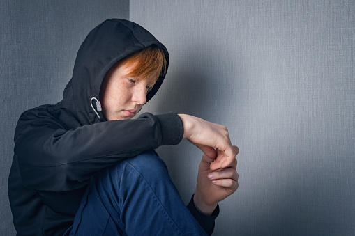 A teenage red-haired boy dressed in a sweatshirt with a hood sits alone at home with his head bowed in a depressed state. Social concept of the problem in adolescence.