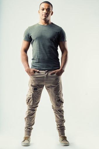 Full length portrait of relaxed macho sexy young Black man in leisurewear standing with legs apart and hands in pockets looking at the camera on white