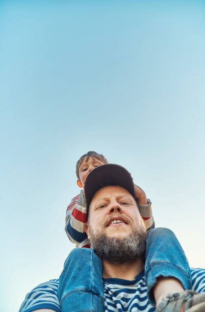 Father with little son sitting on shoulders against blue sky