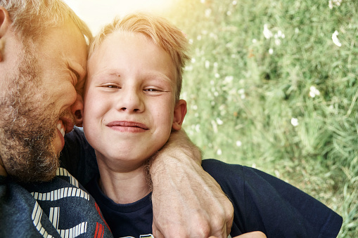 Joyful bearded father hugs handsome smiling blond son spending time in green field with flowers on summer day close view