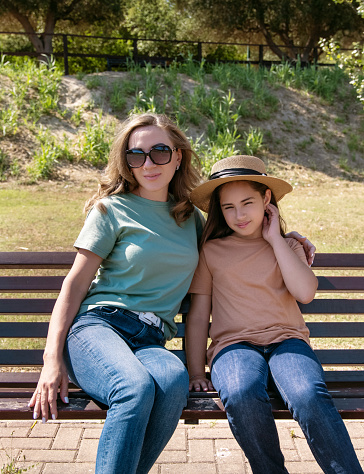 Mother and daughter sitting on the wooden bench in the park in sunny day. A middle-aged woman and kid girl wearing shirts and jeans. T-shirts mockup. Family spending time together in summer