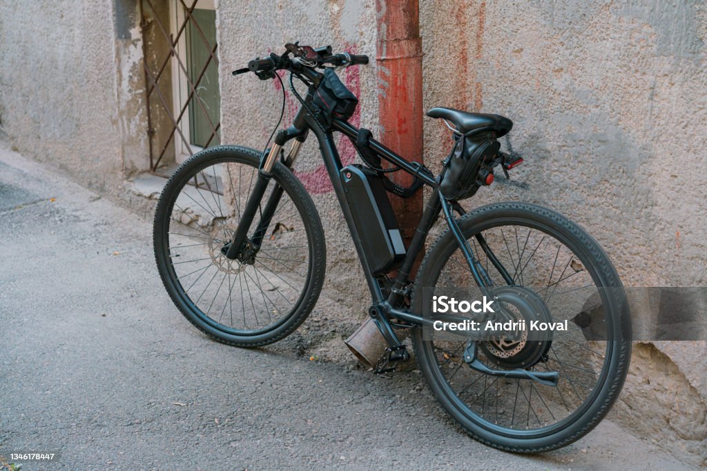 Black electric bicycle with battery on the frame stands on the city's street near the wall. Outdoor. Transportation. Electricity. Electric. Ecological. Electric drive. Engine Electric Bicycle Stock Photo