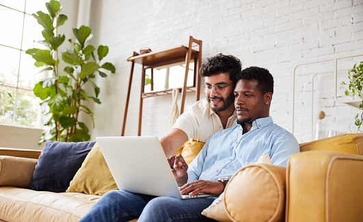 Smiling young multiracial gay couple working online together with a laptop while relaxing in their living room at home