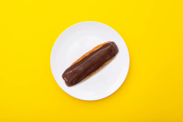 Chocolate eclair on the ceramic plate with bright yellow surface. Minimal composition. Top view.