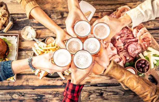 Festive friends enjoying happy hour drinking beer at brewery pub bar restaurant - Young people hands toasting home brew ale dining together - Top view - Party and friendship concept stock photo