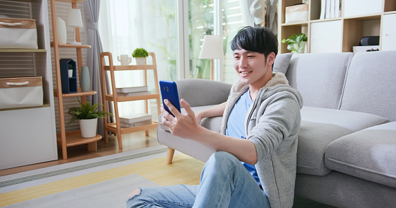 asian young man sitting on floor has video chat by smartphone with friend happily in living room at home