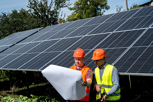 near the new solar panels, two young workers in uniform stand with a plan of work performed. Green electricity concept