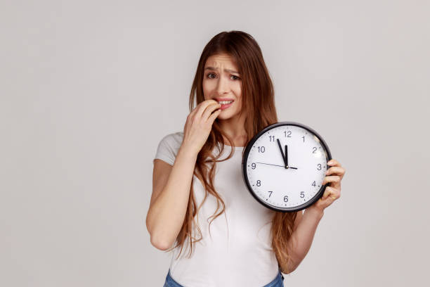 Portrait of nervous dark haired woman biting nails holding big wall clock, deadline, need hurry up. Portrait of nervous dark haired woman biting nails holding big wall clock, deadline, need hurry up, wearing white casual style T-shirt. Indoor studio shot isolated on gray background. time danger stock pictures, royalty-free photos & images