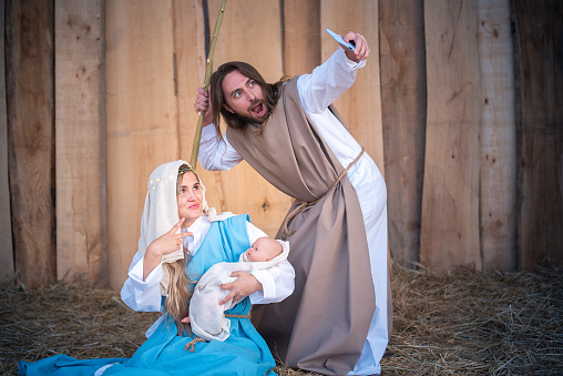 Biblical characters of the virgin mary and joseph taking a selfie while gesturing victory in a crib