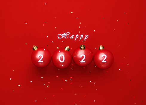 Happy 2022 written red Christmas bauble pair sitting beneath falling confetti over red background. Christmas concept. Horizontal composition copy space. Directly above.