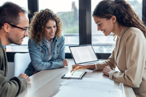 Beautiful real-estate agent showing house plans on electronic tablet while talking to the couple about buying the house in a real estate