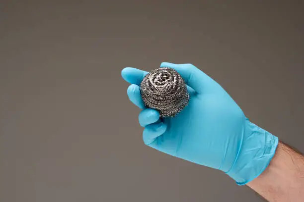 New kitchen metal wire scrubber held in hand by Caucasian male hand wearing a blue latex glove. Close up studio shot, isolated on gray background.