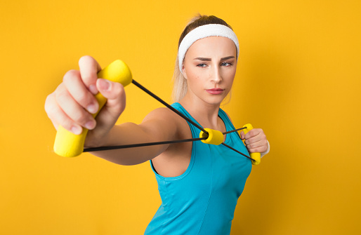 Lazy woman performs exercises for muscles hands with resistance band, isolated on yellow background. Half length of fit sportswoman exercising with expander. Strength motivation. 80s styling. Studio shot