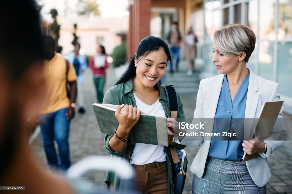 University teacher talking to her Asian female student at campus. Happy Asian student communicating with her professor in front of university building. Professor Stock Photo