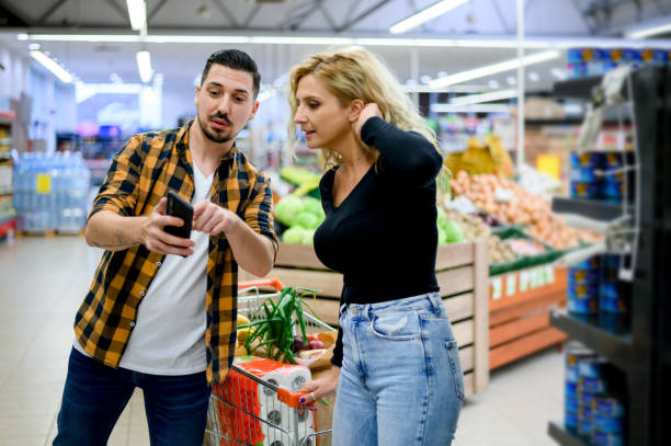 Couple in supermarket using smartphone with shopping cart while grocery shopping Young happy couple using smartphone in supermarket with shopping cart choosing products while grocery shopping supermarket family retail cable car stock pictures, royalty-free photos & images