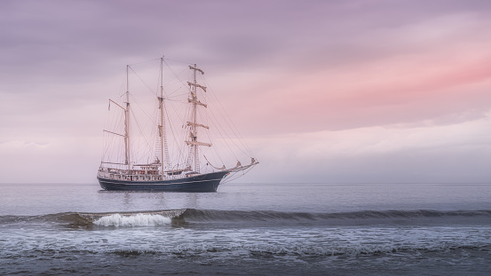 Rolling wave in front of beautiful tall ship covered in haze with soft morning sky, anchored near Northern Ireland coast, Ballycastle