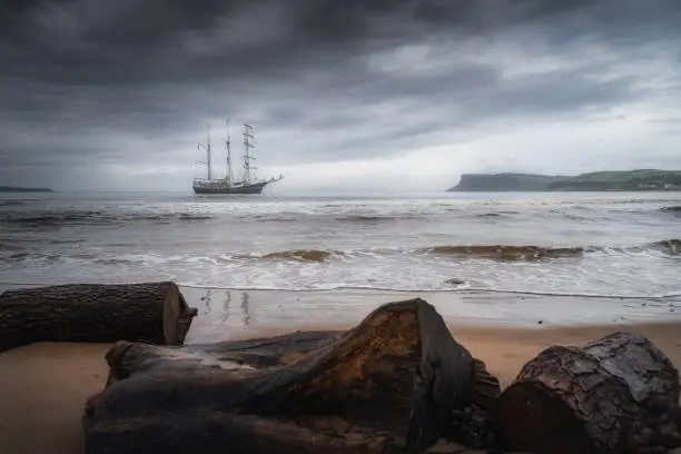Dramatic storm sky rolling over anchored tall ship near Northern Ireland coast, Fair Head in far distance, seen from behind of drift wood on a beach