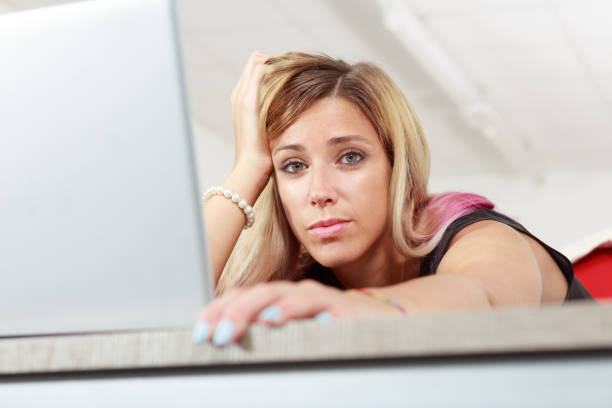Bored businesswoman working on a laptop computer leaning forwards over the keyboard with head a on hand staring down at the camera