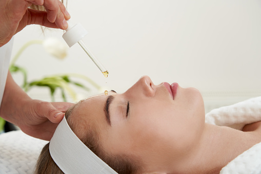 Face Skin Care. Closeup Of Beautiful Smiling Woman Receiving Serum Oil Treatment In Beauty Salon. Cosmetologist Applying Cosmetic Serum On female Face in Cosmetology Center