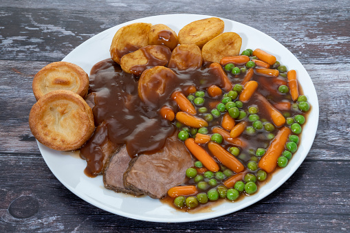 Roast beef dinner in gravy with peas, carrots, roast potatoes, and Yorkshire puddings