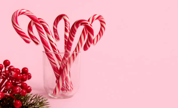 istock Studio shot of beautiful minimalistic simple composition with Christmas lollipops in transparent glass and snowy branch of pine with red berries, holly, on pink background with copy space for ad 1346237242
