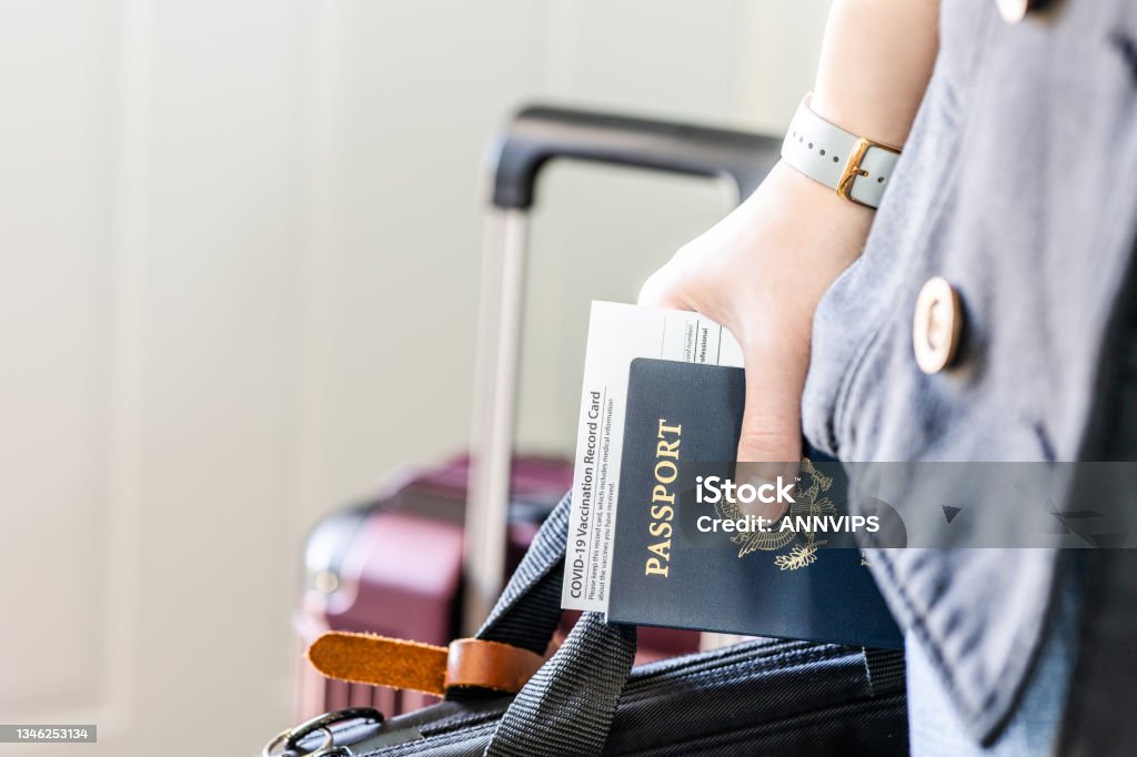 A person with US citizenship identification, vaccinated certified passport and personal belonging ready for travel in new normal. Hand holding passport and approved vaccination card with laptop bag ready for business trip. Vaccine and citizen identity documents of passenger require for travel in new normal. Passport Stock Photo