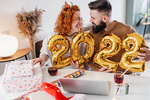 A young couple is at home, they are spending time together during the winter holidays while communicating on a video call. They holing ballon numbers 2022