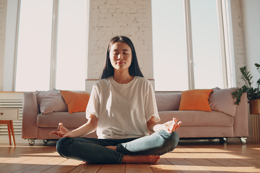 Asian woman doing yoga and zen like meditation lotus pose in casual wear at indoor living room apartment with natural sun light.
