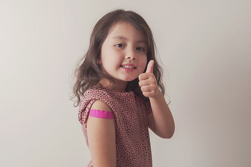 Young mixed Asian girl giving thumb up and showing her arm with pink bandage after got vaccinated or inoculation, child under 12 years old immunization, health concept
