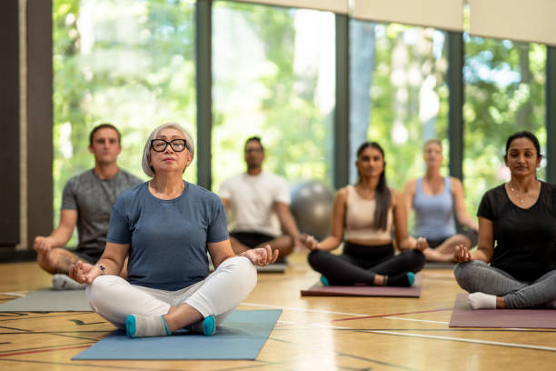 Group Meditation A diverse group of adults meditate on their yoga mats in a gymnasium. There are large windows looking out on a forest in the background. community health center stock pictures, royalty-free photos & images