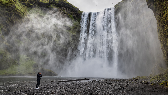 Young man taking photos of Skogafoss Waterfall. Photographer exploring Iceland, taking shots with his camera of the majestic Skogafoss Waterfall in the water spray under moody overcast cloudscape in summer. Panorama Photo. Skogafoss Waterfall, Skógar, Rangárþing eystra, Southern Iceland, Sudurland, Iceland, Nordic Countries, Europe