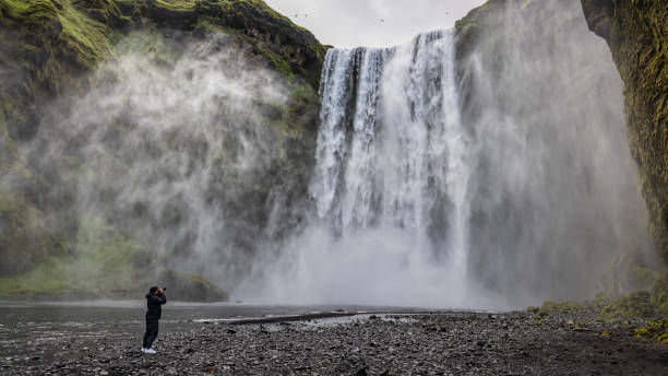 Young man taking photos of Skogafoss Waterfall Skógafoss Iceland Young man taking photos of Skogafoss Waterfall. Photographer exploring Iceland, taking shots with his camera of the majestic Skogafoss Waterfall in the water spray under moody overcast cloudscape in summer. Panorama Photo. Skogafoss Waterfall, Skógar, Rangárþing eystra, Southern Iceland, Sudurland, Iceland, Nordic Countries, Europe national road stock pictures, royalty-free photos & images