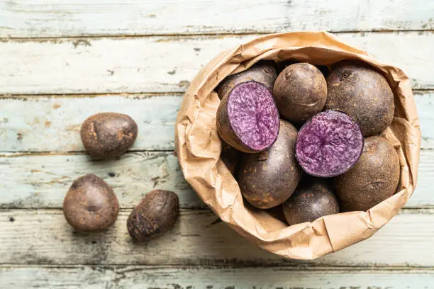 Close up of farm fresh purple potatoes in a paper bag on white wooden background, top view