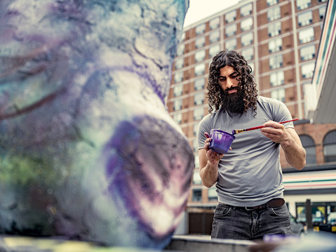 Young artist painting finish on his street sculpture. He has long black curly hair and beard, dressed in t-shirt and jeans. Exterior of city street sidewalk  in downtown district of North American city.