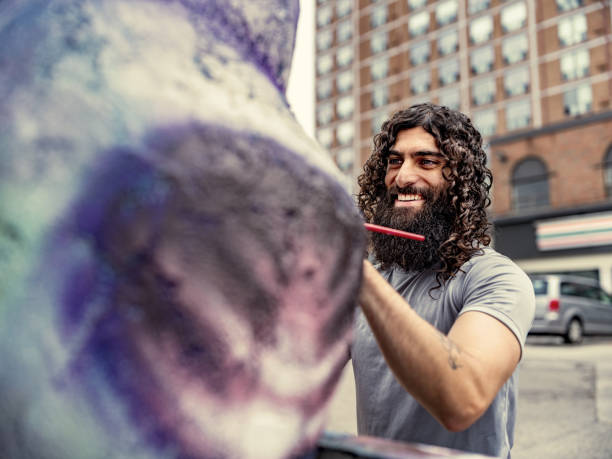 Young artist painting finish on his street sculpture. He has long black curly hair and beard, dressed in t-shirt and jeans. Exterior of city street sidewalk  in downtown district of North American city.