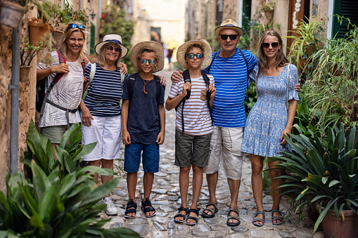 Multi generation family sightseeing beautiful town of Valldemossa. Sunny summer day in Majorca, Spain.\nMother, grandparents and teenagers are walking in narrow street decorated with potted plants.\nCanon R5