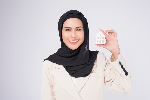 Young beautiful muslim woman in suit holding small model house over white background studio