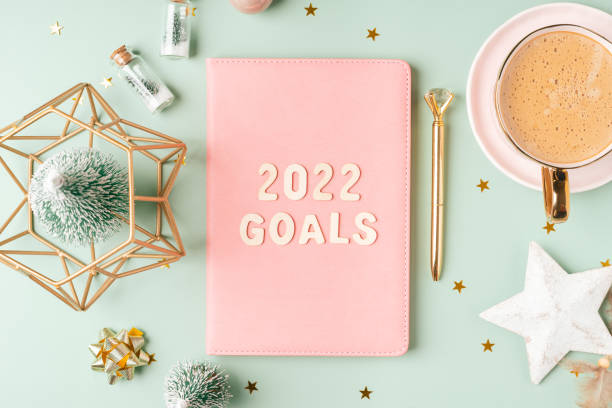 istock Top view Desktop Christmas pink notepad with 2022 goals letters text. Flat lay on green mint table background with planner, cup of coffee, candle, Christmas decoration, notebook and stationery 1346323857