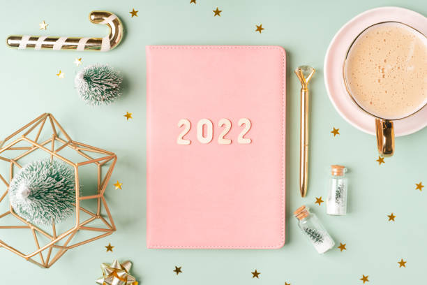 istock Top view Desktop Christmas pink notepad with 2022 letters text. Flat lay on green mint table background with planner, cup of coffee, candle, Christmas decoration, notebook and stationery 1346323922