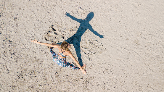 Drone view of woman on the sand standing legs apart, arms outstretched and bending over, her shadow in shape of her figure, doing exercise outdoors