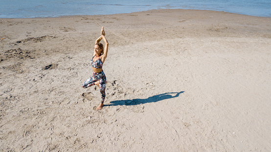 Drone shot of woman exercising on sandy beach, standing on one leg, the other bent inwards, arms raised above head with hands clasped, casting shadow, yoga tree pose practiced outdoors